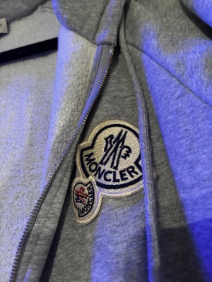 Grå hoodie Moncler - Grå hoodie från Moncler med broderade logopatchar på bröstet, dubbla dragkedjor och fickor med dragkedja. Insidan är fodrad med mjuk fleece och jackan har justerbar huva med snörning. Passar 10/10 till varenda outfit du kan tänka dig, super skön & varm och ha på sig.                        Huvud Dragkedja funkar ej, saknar dragkedja på en ficka