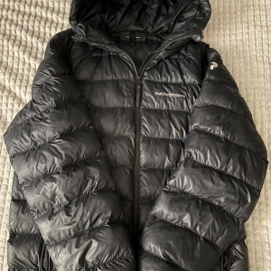  Peak Performance vinterjacka - Svart pufferjacka från Peak Performance med huva och dragkedja framtill. Jackan har vadderade paneler, praktiska stora och fodrade fickor med dragkedja. Perfekt för kalla dagar som är runt hörnet, med en look som passar ”alla” stiler.