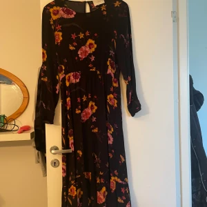 Svart långklänning med blommor från JUST - Svart långklänning från JUST i storlek S med färgglatt blommigt mönster i rosa, gult och lila. Klänningen har långa ärmar och rund halsringning. Materialet känns mjukt och lätt, perfekt för dig som gillar romantiska och färgstarka plagg.