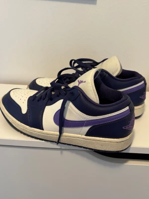Nike Air Jordan 1 Low vit/lila - Original Nike Air Jordan 1 Low sneakers i vit och mörklila med lila detaljer och broderad Air Jordan-logga på hälen och Jumpman-logga på tungan. Har använd mindre än 5 gånger. Nästan helt nya! Kan skicka fler bilder i dm. Storlek EUR 40 (25,5 cm)  men passar 39-40. Priset kan diskuteras.