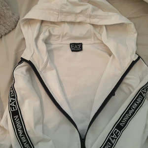 Vit EA7 hoodie med logoband - Snygg vit hoodie från EA7 Emporio Armani med svarta logoband längs ärmarna och dragkedja framtill. Tröjan har huva med snörning och ribbade muddar. Perfekt för dig som gillar stilrena streetwear-plagg med tydlig branding.