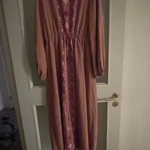 Lång klänning  - Långklänning i dova rosa och beige toner med bohemisk känsla. Klänningen har långa ärmar och ett mönstrat parti framtill med små geometriska former. Den har markerad midja och ett luftigt fall, perfekt för dig som gillar avslappnad och trendig stil.