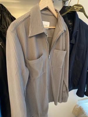 Grå ribbad overshirt från Ciszere - Säljer en stilren grå overshirt från Ciszere med ribbad struktur, två stora bröstfickor och dragkedja framtill. Skjortan har klassisk krage och långa ärmar, perfekt för lager-på-lager-look. Materialet känns mjukt och följsamt. Jag är 1,89 meter lång och men relativt smal, för liten för mig alltså. Den är i storlek M men passar om man har storlek L vanligtvis. 