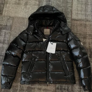 Moncler Maya jacka - Säljer en helt ny Moncler Maya jacka. Svart pufferjacka från Moncler med glansig finish och huva. Jackan har dragkedja framtill, två sidofickor med dragkedja och coolt Moncler-märke på ärmen. Insidan har ett unikt serietryck och flera etiketter. Perfekt för kalla dagar. Alla tags, etiketter, Qr kod samt RFID och NFC Scan som tar dig till Monclers officiella hemsida finns.