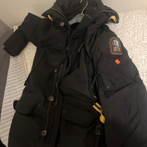 Svart Parajumpers parkajacka XL - Svart parkajacka från Parajumpers i storlek XL med slim fit. Jackan har flera fickor, gul detalj vid dragkedjan, patch på ärmen och robust huva. Materialet är slitstark polyester och jackan har både knappar och dragkedja framtill.