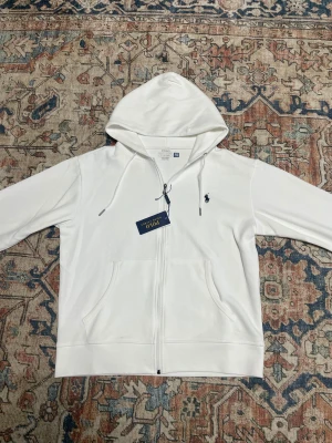 Polo Ralph Lauren - Vit hoodie från Polo Ralph Lauren med klassisk broderad logga på bröstet. Tröjan har huva med snörning, dragkedja framtill och två fickor. Tillverkad i mjuk bomull för skön komfort. Perfekt för chill dagar och enkel att matcha med jeans eller joggers.
