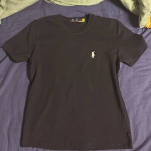 Ralph Lauren T-shirt - Svart Ralph Lauren T-shirt – Storlek L Helt oanvänd Ralph Lauren T-shirt i svart. Klassisk modell i mjuk bomull med den ikoniska broderade loggan på bröstet. Sitter snyggt och funkar till alla outfits. Inga fel eller märken eftersom den aldrig är använd.