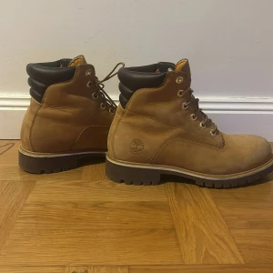 Timberland boots i brunt skinn - Säljer ett par klassiska Timberland boots i brunt skinn med svart vadderad krage och grov sula. De har snörning med metallöglor och rund tå. Perfekta för höst och vinter, och riktigt snygga till jeans eller cargo pants.
