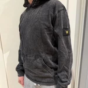 Mörkgrå hoodie från Lyle & Scott - Snygg mörkgrå hoodie från Lyle & Scott Junior med klassisk gul logga på ärmen. Tröjan har huva, ribbade muddar och en stor magficka. Materialet är mjuk bomull och passformen är normal, perfekt för chill dagar. Endast provad så helt i nyskick!