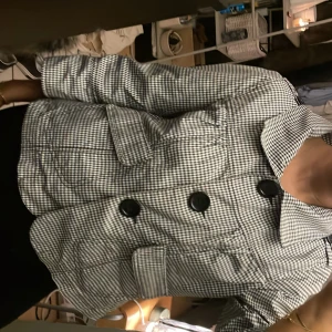 Svartvit rutig kavaj med stora knappar - trekvarts - Snygg svartvit rutig kavaj med stora svarta knappar och bred krage. Jackan har två framfickor med lock och en kortare, boxig passform. Storlek L men sitter lite oversized på en S