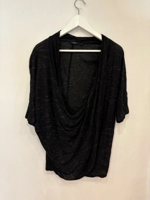 Svart draperad topp från Lindex - Svart topp från Lindex med draperad, djup v-ringning och korta ärmar. Tunt, lätt material med diskreta gråa ränder som ger en cool effekt. Oversized passform som faller snyggt och känns riktigt chill.