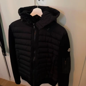 Svart Moncler Cardigan - Svart dunjacka från Moncler med quiltad front och huva. Jackan har stickade ärmar, dragkedja framtill och en ficka med dragkedja på ärmen. Storlek M och den är som ny. Använd ca 2 gånger. Skriv till mig.