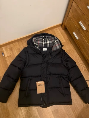 Svart pufferjacka från Burberry - Svart pufferjacka från Burberry med huva och klassiskt rutig foder i grått, vitt och rött. Jackan har dragkedja och knappar framtill, två fickor med dragkedja och en patch med Burberry London England på ärmen. Perfekt för kalla dagar.