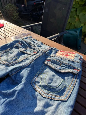 True Religion jeans vintage med teal stitch - Säljer ett par äldre blå jeans från True Religion med broderade bakfickor och kontrastsömmar. Jeansen har knappar med logga, detaljer i rött och en patch med varumärket bak. Materialet är klassisk jeans och passformen är loose/straight fit. De säger W34 men sitter lite lösare och benen är 33 ish skulle jag säga men sitter ändå fett najs pga det är straight fit