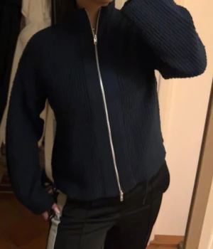 Mörkblå stickad kofta med dragkedja - Säljer en mörkblå stickad kofta med hel dragkedja framtill och hög krage. Koftan har långärmade och en ribbad struktur som ger en snygg och modern look. Perfekt att slänga på sig när det är lite kyligt ute.