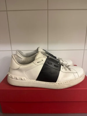 Valentino Garavani Open sneakers vit/svart - Säljer ett par snygga Valentino Garavani Open sneakers i vitt skinn med bred svart läderpanel över snörningen och coola nitar på hälen. Klassisk rund tå och platt sula, med originalskokartong och extra skosnören. Perfekt för dig som gillar lyxiga street vibes. Köpte på Vestiaire. 