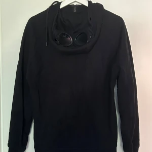 Svart hoodie med goggles från C.P. Company - Svart hoodie från C.P. Company med ikoniska goggles i huvan och dragkedja framtill. Tröjan har två snedställda fickor med dragkedja och långa ärmar. Perfekt för dig som gillar streetwear och vill sticka ut med en unik detalj.