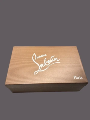 Grå Christian Louboutin sneakers med nitar - Säljer ett par grå sneakers från Christian Louboutin med coola nitar på tån och klassisk röd sula. Skorna har snörning och är gjorda i mocka med en rund tå. Insidan är beige och de har en platt sula. Perfekt för dig som vill sticka ut med lyxig streetstyle.