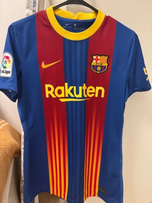 Barça matchtröja Nike blå/röd - Barcelona matchtröja från Nike med klassiska blå och röda vertikala ränder, gula detaljer och klubbmärke på bröstet. La Liga-patch på ärmen och sponsorer som Rakuten och Beko tryckta i gult. Tillverkad i lätt polyester, perfekt för fotboll.
