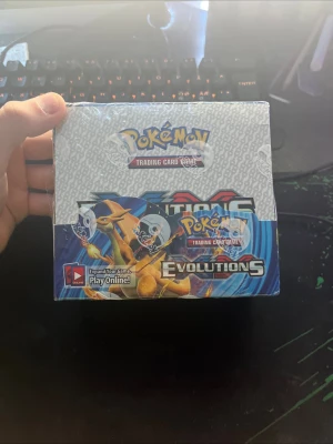 Pokémon Evolutions boosterbox - Säljer en Pokémon TCG Evolutions boosterbox med coola motiv av Charizard, Blastoise, Raichu och Venusaur på förpackningen. Boxen har en färgstark design i blått, orange och grått med Pokémon-logga och Evolutions-tryck. Perfekt för dig som vill utöka din samling eller spela online.
