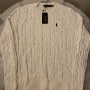 Vit Polo Ralph Lauren stickad tröja - Vit kabelstickad tröja från Polo Ralph Lauren med klassisk rund halsringning och broderad marinblå logga på bröstet. Tröjan har ribbade muddar och är tillverkad i mjuk bomull. Perfekt för en clean och stilren look.