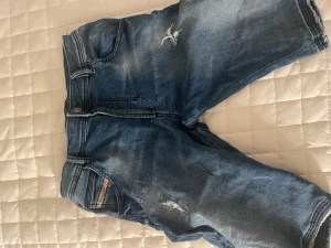 Blå denimshorts från Dsquared2 - Säljer ett par blå denimshorts från Dsquared2 med slitna detaljer och klassisk femficksdesign. Shortsen har knapp och dragkedja framtill samt en liten logotyp vid fickan. Perfekta för en chill och avslappnad stil.