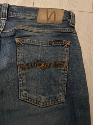 Blå jeans från Nudie Jeans, W30 L34 - Säljer ett par klassiska blå jeans från Nudie Jeans med snygga orangea sömmar och vågig brodyr på bakfickorna. Modellen har normal passform och raka ben, tillverkade i slitstarkt bomullsmaterial. Perfekt för dig som gillar stilrena och tidlösa jeans.