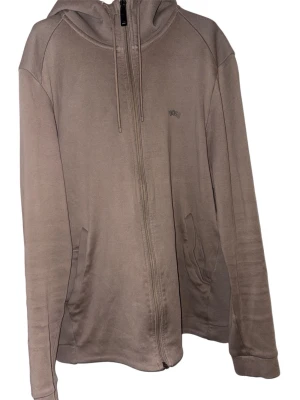 BOSS beige hoodie,  - En ljusbrun hoodie med dragkedja från BOSS. Den har en huva med dragsko och två fickor fram. Logotypen är diskret placerad på bröstet.