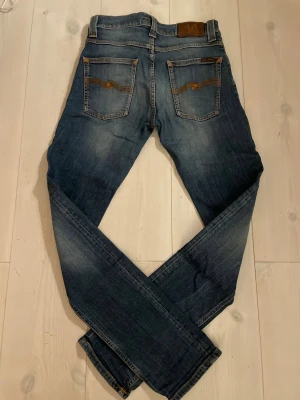 Blå Nudie Jeans  - Snygga blå jeans från Nudie Jeans med slim fit  och klassisk femficksdesign. Jeansen har gul kontrastsöm och Nudie-logga på bakfickan. Materialet är slitstarkt denim i bomull och passformen är tight hela vägen ner. Kan vara lite slitage men det har jag inte märkt alls under tiden jag haft på mig dem. Perfekt för dig som gillar en schysst look!