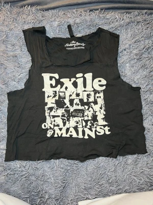 The Rolling Stones grå croppad linnetopp - Grå croppad linnetopp från The Rolling Stones med vit tryckt grafik och text 'Exile on Main St' på framsidan. Toppen är ärmlös och har råa kanter för en edgy vibe. Materialet är mjuk bomull och passformen är loose. Perfekt för dig som gillar bandmerch och streetstyle.