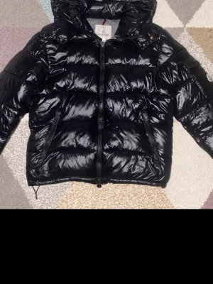 Svart Moncler pufferjacka med huva -  Moncler maya  glansig finish och huva. Jackan har dragkedja framtill, två sidofickor med dragkedja och ett grått innerfoder. Klassisk Moncler-logga på ärmen. Helt äkta och nfc funkar som det ska 