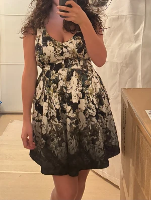 Blommig satinklänning med v-ringning - Blommig satinklänning med blommigt mönster i vitt och grönt. Klänningen har v-ringning och breda axelband, markerad midja och utställd kjol. Finns två fickor. I mycket fint skick. 