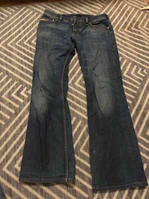 Diesel bootcut jeans blå - Snygga blå jeans från Diesel, med klassisk bootcut och raka ben. Jeansen har detaljerade sömmar och logga på bakfickan. Tillverkade i slitstarkt denim med knappgylf och coola nitar med Diesel-logga. Jeansen är i bra skick men har hål på insidan, men dem syns inte från utsidan!