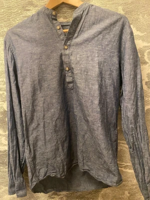 Blå henley från Jack & Jones - Säljer en blå henley-skjorta från Jack & Jones Essentials, tillverkad i en skön mix av bomull och linne. Skjortan har lång ärm, halv knäppning med fyra knappar och en enkel, stilren design. Perfekt för dig som gillar avslappnad och modern stil.