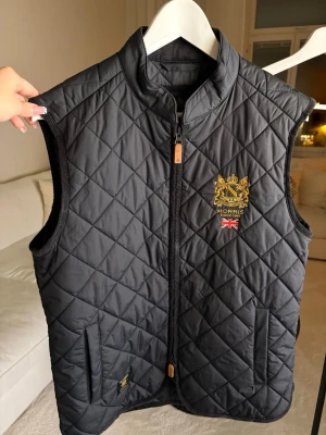 Svart quiltad väst från Morris - Quiltad svart väst från Morris med guldbroderad logga och brittisk flagga på bröstet. Västen har ståkrage, två fickor och dragkedja framtill. Snyggt rutmönster och exklusiv känsla, perfekt för dig som gillar klassisk stil.