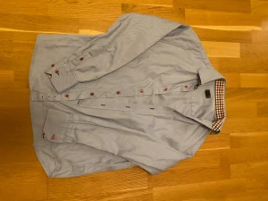 Blårandig skjorta från H&M - Snygg blårandig skjorta från H&M med röda knappar och rutigt tyg på insidan av kragen. Skjortan har lång ärm och klassisk krage, perfekt för dig som gillar stilrena detaljer. Materialet är mjuk bomull som känns skönt mot huden.