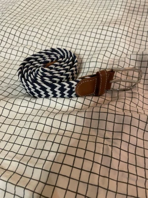 Mörkblå och vit flätad bälte - Säljer ett snyggt flätat bälte i mörkblått och vitt med bruna skinn-detaljer och silvrigt spänne. Bältet har ett zigzag-mönster och passar perfekt till jeans eller chinos. Stilren och modern look som lyfter outfiten.