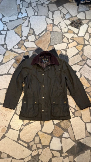 Barbour olivgrön parkajacka - Klassisk olivgrön parkajacka från Barbour med bruna manchesterdetaljer på kragen och rutigt innerfoder. Jackan har stora fickor med tryckknappar och en rak passform. Perfekt för dig som gillar tidlös stil och funktion.