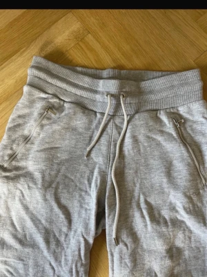 Grå mjukisbyxor från Röhnisch M - Snygga grå mjukisbyxor från Röhnisch i storlek M. Byxorna har ribbad midja med snörning, dragkedjor på fickorna och muddar vid bensluten. Tillverkade i mjukt bomullsmaterial som är perfekt för chill eller träning. Enkla att matcha med hoodies eller t-shirts.