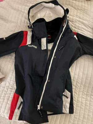 Marinblå vindjacka från Marine - Snygg marinblå vindjacka från Marine med vita och röda detaljer. Jackan har huva, dragkedja, meshfoder och praktiska fickor. Perfekt för blåsiga dagar och outdoor-aktiviteter. Sportig jacka och väldigt skön!