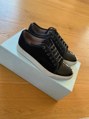  Lanvin skor svarta  - Snygga svarta sneakers från Lanvin med vit platt sula och snörning. Skorna har en blank tå i lack och resten är i mocka, vilket ger en cool kontrast. Perfekta för dig som gillar stilrena och exklusiva sneakers med lite edge.