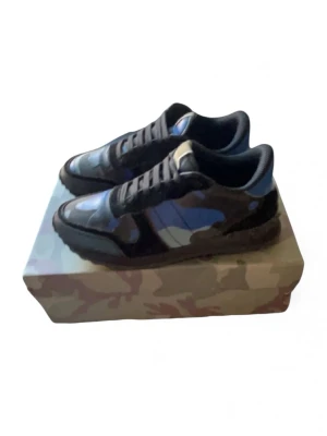 Valentino Rockrunner sneakers med blått camomönster - Säljer ett par Valentino Rockrunner sneakers med blått och svart camouflage-mönster. Skorna har snörning, rund tå och platt sula. Materialet är en mix av skinn och textil, vilket ger en lyxig känsla. Perfekta för dig som vill sticka ut med en unik design.