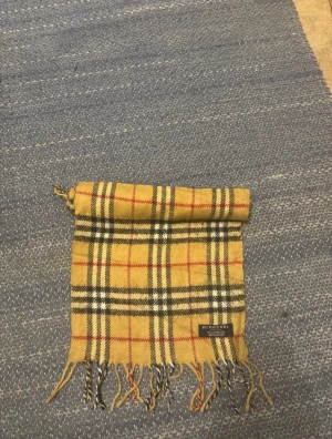 Rutig beige Burberry halsduk - Säljer en klassisk beige halsduk från Burberry med det ikoniska rutiga mönstret i svart, vitt och rött. Halsduken är tillverkad i 100% lammull och har fransar i båda ändar. Perfekt för att ge din outfit en lyxig touch.