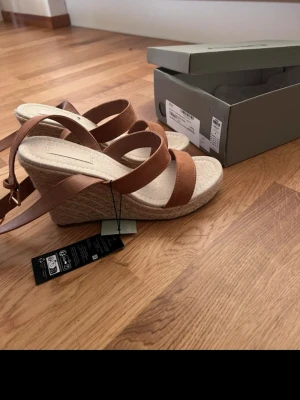 Bruna wedge sandaler från ONLY, strl 40 - Säljer ett par snygga wedge sandaler från ONLY i storlek 40. Sandalerna har bruna remmar och en beige flätad kilklack. De har öppen tå och spänne vid ankeln. Perfekta för sommarens alla tillfällen och riktigt sköna att gå i.