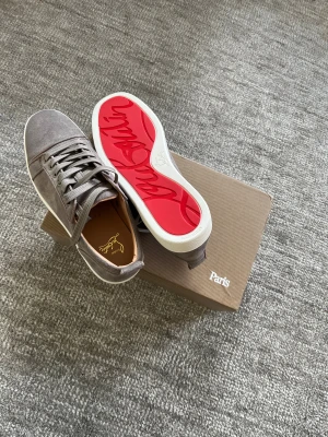 Gråa Christian Louboutins - Säljer ett par gråa sneakers från Christian Louboutin med röd sula. Skorna har en rund tå och är tillverkade i mocka med vita detaljer på sulan. Kommer med originalkartong, dustbag och äkthetskort. Perfekt för dig som gillar lyxiga detaljer. Modellen utan spikar. Skorna är inte så mycket använda, men sulan på ena skon är lite uppskrapad. Skriv om fler bilder behövs