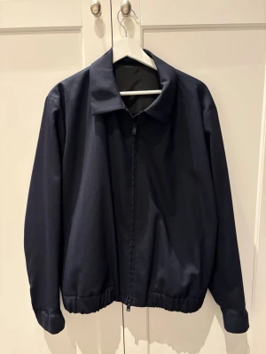 Mörkblå bomberjacka från Massimo Dutti - Snygg mörkblå bomberjacka från Massimo Dutti med klassisk krage och dragkedja framtill. Jackan har elastisk nederkant och är tillverkad i ett stretchigt syntetmaterial som är lättskött och anti-crease. Perfekt för dig som gillar stilren och enkel design.