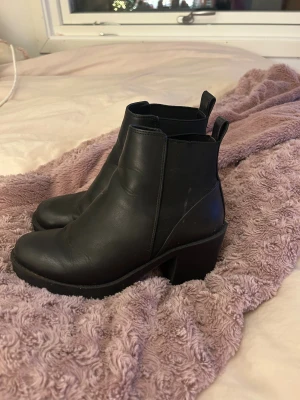 Svarta chelsea boots med klack - Säljer ett par svarta chelsea boots med rund tå och rejäl klack. Skorna är i skinn och har elastiska paneler på sidorna för enkel påtagning. Perfekt för dig som gillar stilrena och klassiska boots med lite höjd.