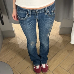 Rock Revival bootcut jeans  - Säljer ett par blå bootcut jeans från Rock Revival med snygga slitningar och detaljerade bakfickor med broderi och metallknappar. Jeansen har låg midja, klassisk femficksdesign och grova kontrastsömmar. Jag säljer dom då dom inte kommer till användning. Man kan se på sista bilden att dom har ett litet hål vid fickan. Även små slitningar längst ner på slutat av byxorna, annars ett bra skick!Skriv bara om fler frågor!❤️❤️❤️