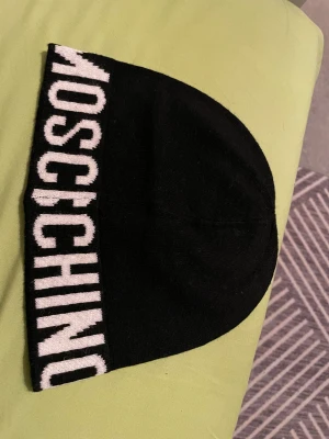 Svart Moschino mössa med logga - Svart mössa från Moschino med bred vit logotyp runt kanten. Stilren och enkel design med mjukt stickat material som sitter skönt på huvudet. Perfekt för dig som vill ha en snygg accessoar med tydlig branding. Mössan är i nytt skick!