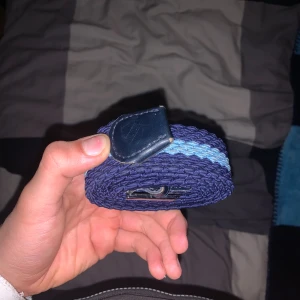 Mörkblå flätad bälte med ljusblå detalj - Snyggt mörkblått flätat bälte från Djurgården  med en ljusblå rand längs mitten. Bältet har ett stilrent spänne i metall och avslut i mörkblått skinn. Perfekt för att ge outfiten en cool touch och funkar till både jeans och chinos.